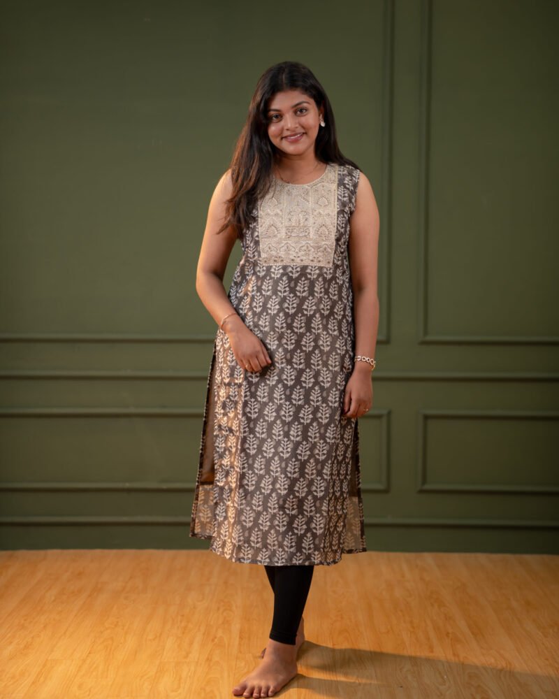 Woman wearing an olive grey A-line kurti with embroidered yoke and subtle leaf print, paired with black leggings, standing against a green panelled background.