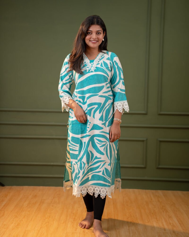 Model wearing a teal blue abstract printed kurti with lace detailing on the sleeves and hem, paired with black leggings, standing against a green paneled background.