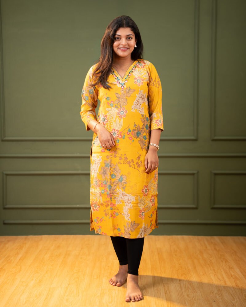 Woman wearing a mustard yellow floral print straight fit kurti with a V-neck and three-quarter sleeves, paired with black leggings, standing against a neutral background.