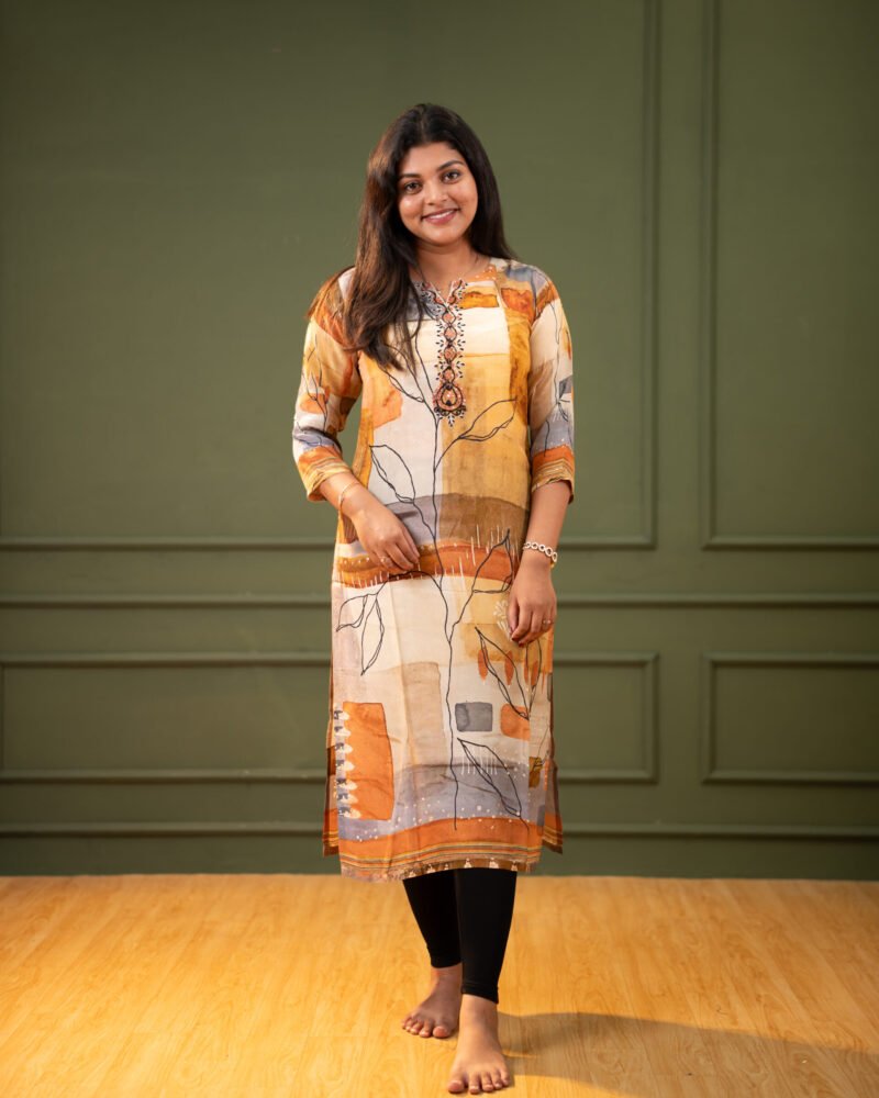 A woman wearing a knee-length straight-fit kurti with a mustard, beige, and rust abstract geometric print and black leaf outlines, standing against a dark green wall.